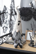 Seated Halloween gnome with a white ghost-face hat, gray beard, striped legs and black shoes in a tabletop display.