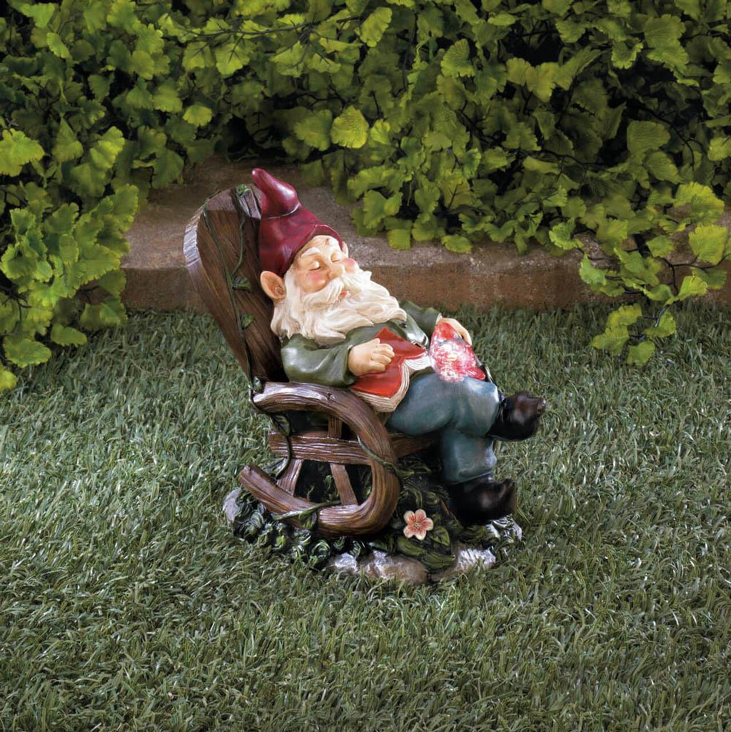 Red-hatted gnome reclining in a wooden-style rocking chair with a book on its lap, displayed on grass.