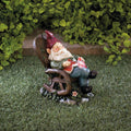 Red-hatted gnome reclining in a wooden-style rocking chair with a book on its lap, displayed on grass.