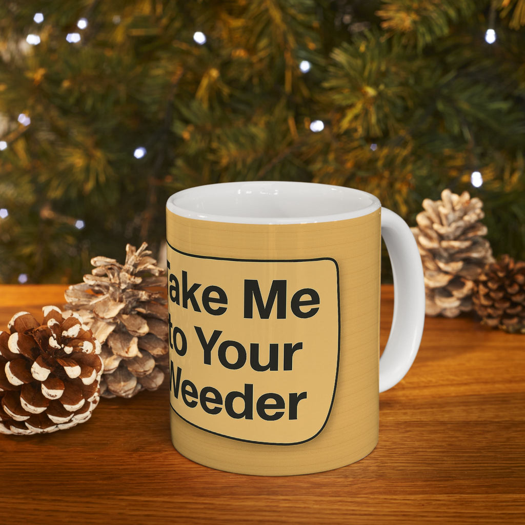Yellow mug with 'Make Me Your Weeder' text in front of a Christmas tree and pinecones.