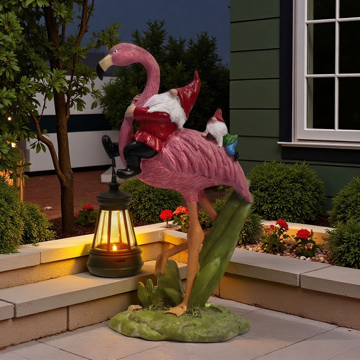 Pink flamingo garden figure carrying two red-hatted gnomes and a glowing lantern, shown on a patio at night.
