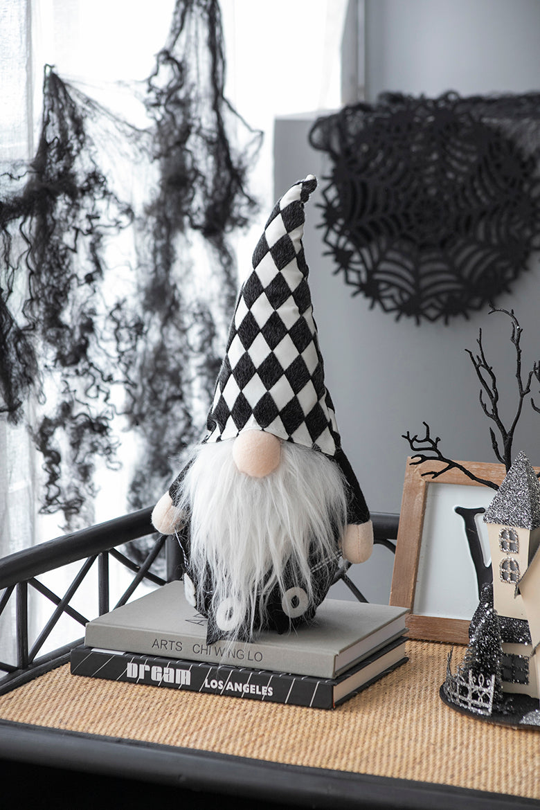 Halloween gnome with a black-and-white diamond-patterned hat and long white beard, sitting on books beside seasonal decor.