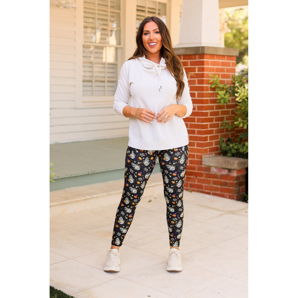Model wearing black leggings with a colorful Halloween gnome pattern, a white hooded top and white sneakers outdoors.