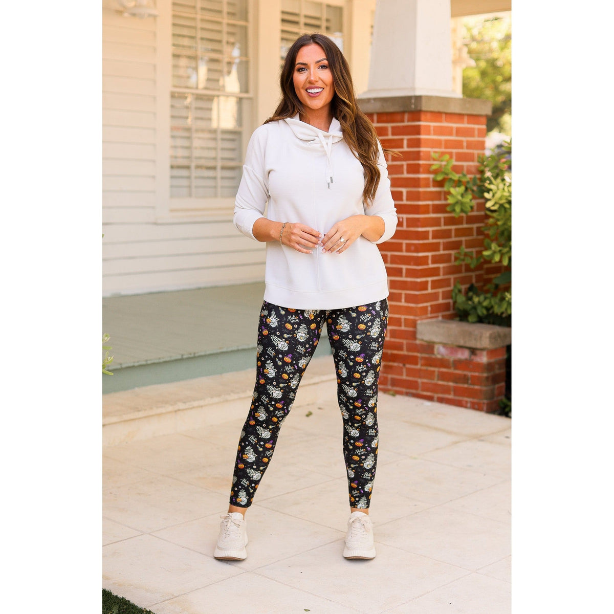 Model wearing black leggings with a colorful Halloween gnome pattern, a white hooded top and white sneakers outdoors.