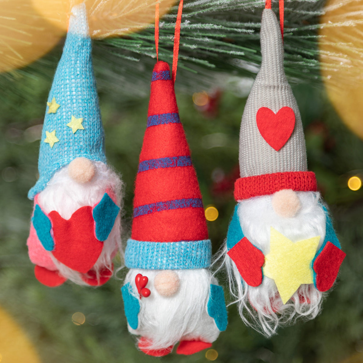 Three hanging fabric gnome ornaments with blue, red and gray hats, white beards, and heart and star accents.