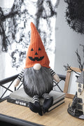 Seated Halloween gnome with an orange jack-o'-lantern hat, gray beard and black-and-white striped arms and legs.
