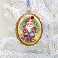 Oval Christmas ornament with a gold-colored edge depicting a red-hatted gnome holding holly and a bell.