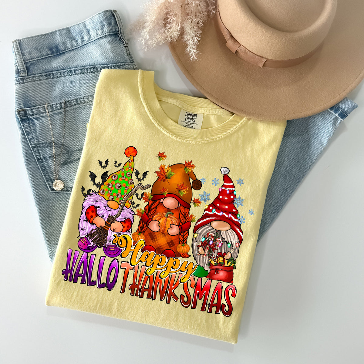 Yellow shirt with Halloween, autumn and Christmas gnomes above Happy HalloThanksMas lettering, shown with jeans and a hat.