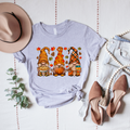 Light gray shirt printed with three autumn gnomes holding a pie, pumpkin and cup, surrounded by leaves and pinecones.
