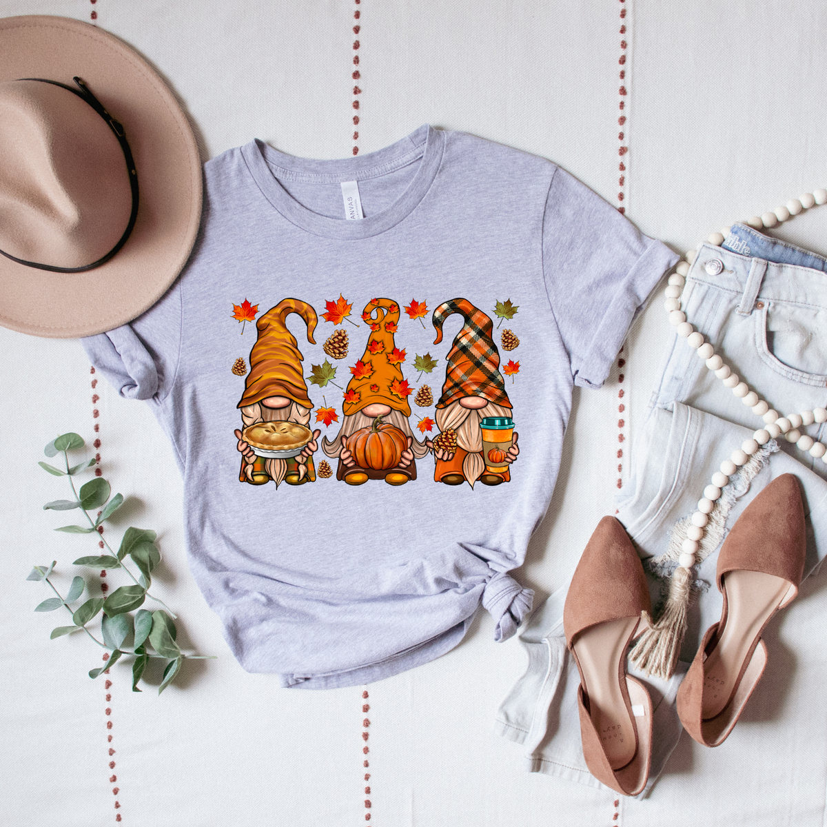 Light gray shirt printed with three autumn gnomes holding a pie, pumpkin and cup, surrounded by leaves and pinecones.