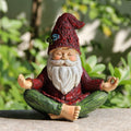 Cross-legged meditating gnome with closed eyes, red hat and coat, green trousers and palms raised.