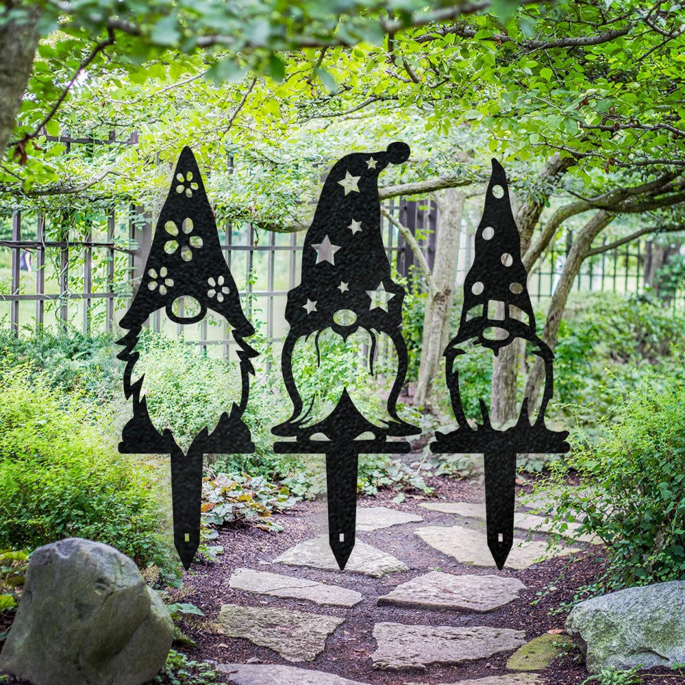 Three black metal gnome yard stakes with flower, star and polka-dot hat cutouts beside a garden path.