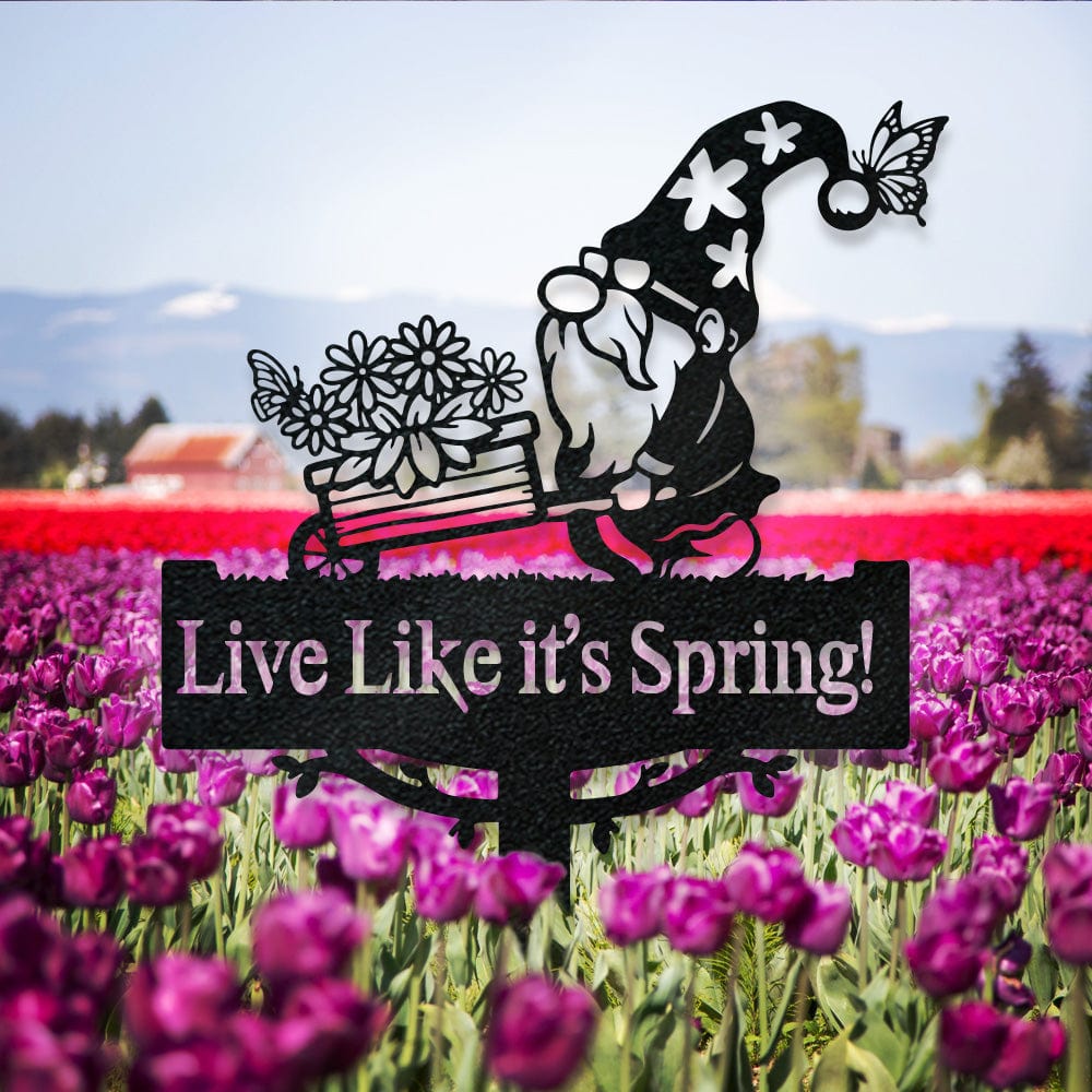 Black metal gnome pushing a flower-filled wheelbarrow above a sign reading Live Like it's Spring! among tulips.