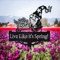 Black metal gnome pushing a flower-filled wheelbarrow above a sign reading Live Like it's Spring! among tulips.