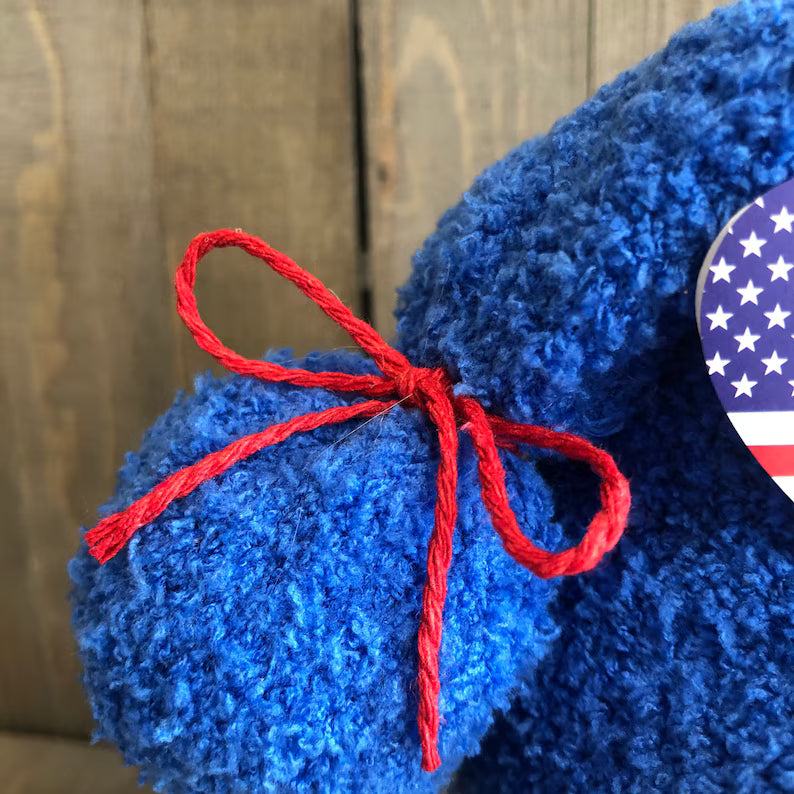 Blue teddy bear with a red bow and American flag patch on a wooden background
