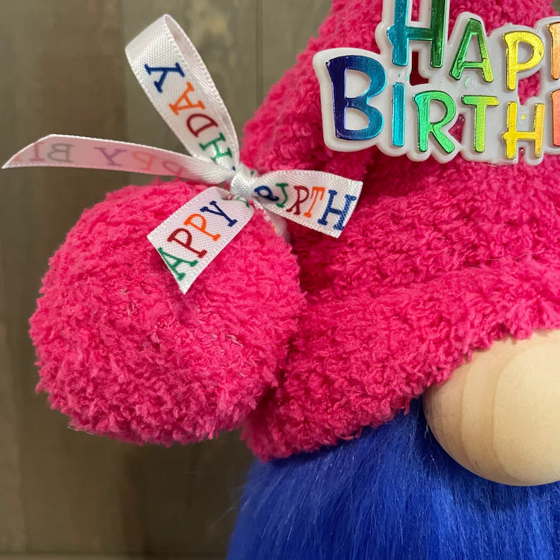 Gnome with pink hat and blue hair wearing a 'Happy Birthday' ribbon against a neutral background